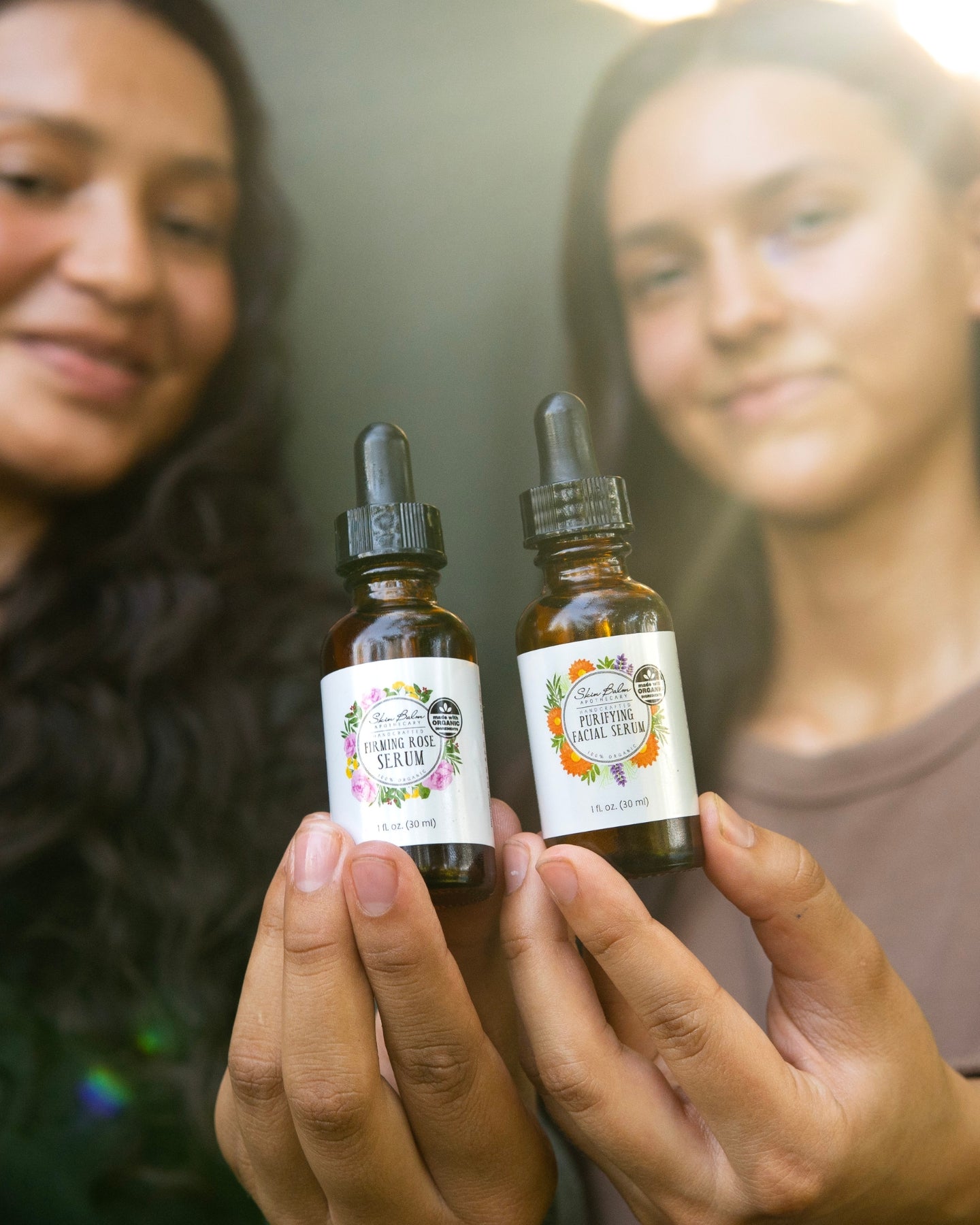 A close-up shot of the Firming Serum and the Purifying Serum being held side-by-side, with the two women holding the serums blurred in the background.