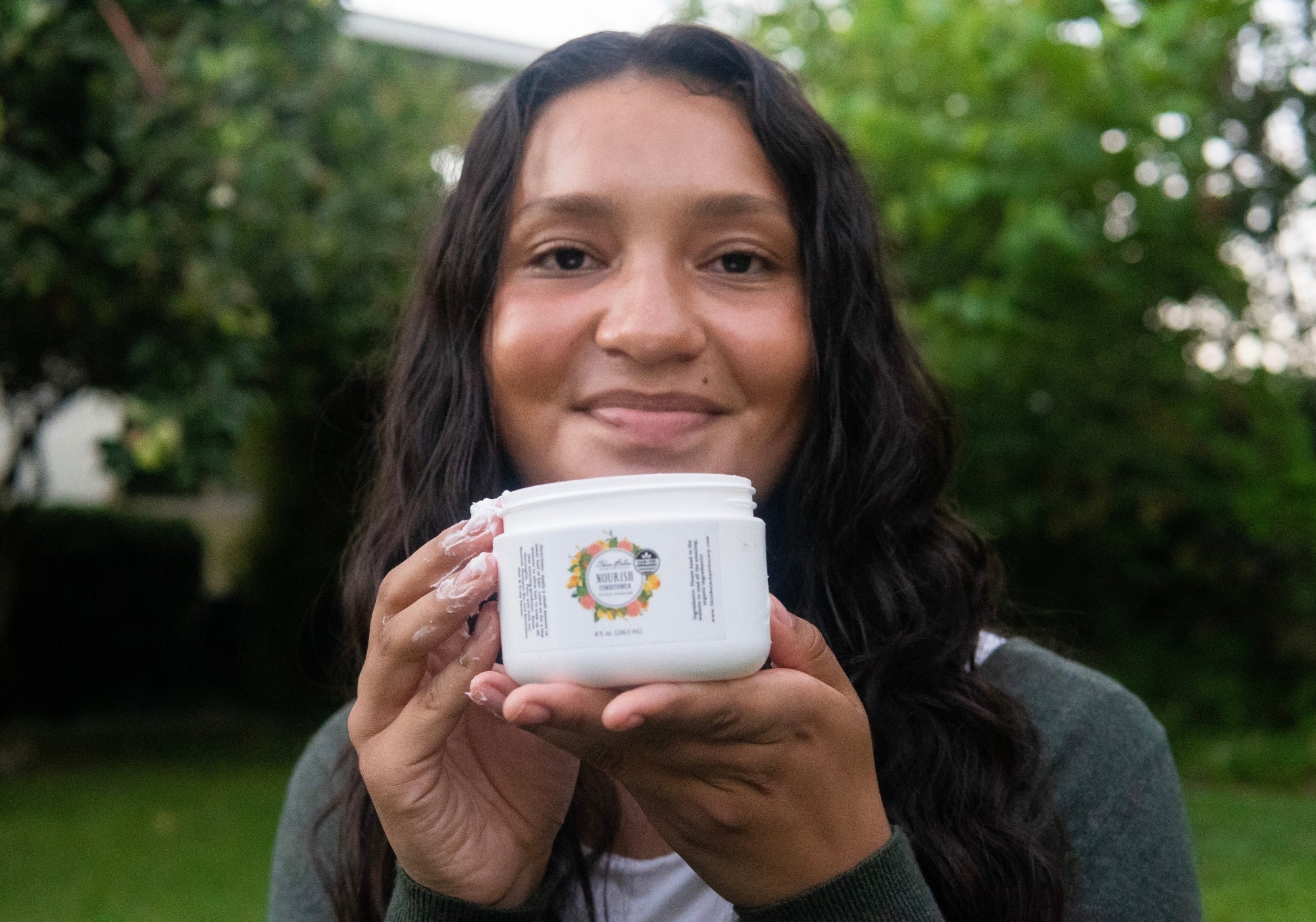 A smiling woman with Nourish Conditioner on her fingers holds the container to her face.