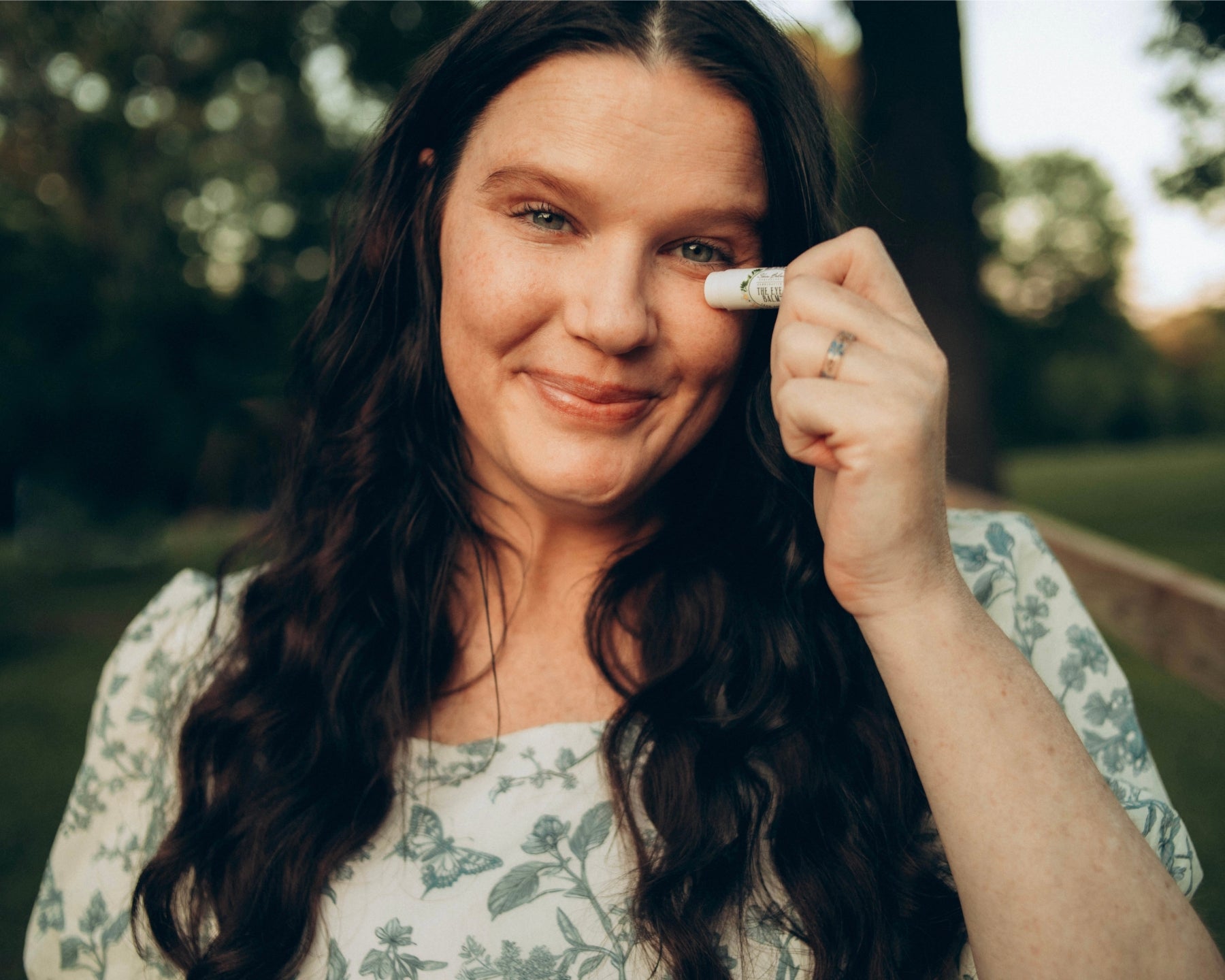 A smiling woman in a powder blue, floral print dress applies The Eye Balm™.